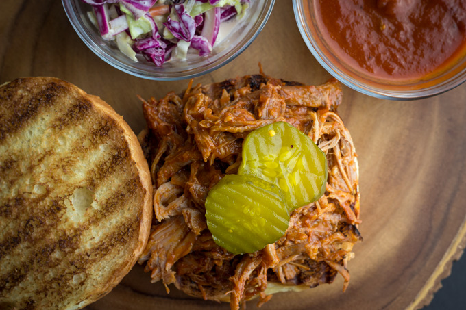 Top view of an open BBQ Pork sandwich with pickles and a bowl of onions and bbq sauce at the sides